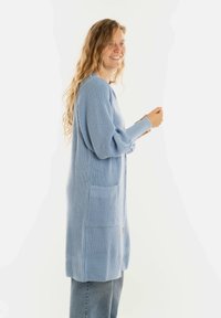 Jonge vrouw met lang, krullend haar, gekleed in een lichtblauwe gebreide cardigan en spijkerbroek, glimlachend en naar de zijkant kijkend.