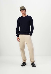 Tengerészkék kötött pulóver, bézs nadrág és fekete cipő. A sapka kockás mintás. A modell egyszerű háttér előtt áll.