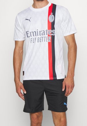 Mann trägt ein weißes AC Milan Fußballtrikot mit rot-schwarzen Streifen und schwarzen Puma-Shorts vor einfarbigem Hintergrund.