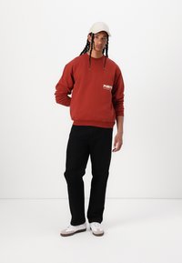 Rød bomullsgensar med "POMPEI" logo på brystet, svarte bukser og hvite joggesko. Modellen har på seg en beige caps og har flette.