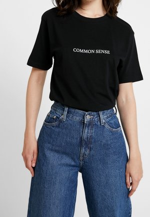 Osoba w czarnej koszulce z napisem "COMMON SENSE" i wysokim stanem niebieskich dżinsów, stojąca na tle białej ściany.