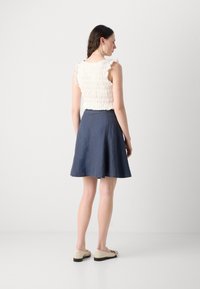Light pink smocked top with ruffled sleeves, paired with a navy flared skirt and beige shoes with black soles. Rear view.