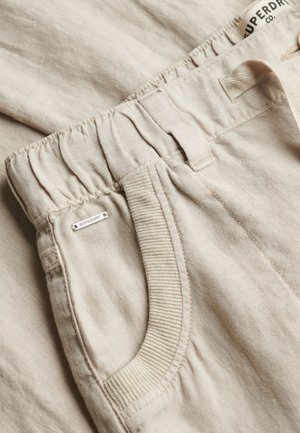 Beige linnen broek met elastische tailleband, geribbelde zakafwerking, broekriemlussen en een klein metalen Superdry-logolabel bij de zak.