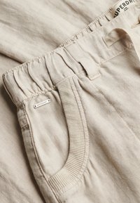 Pantalon en lin beige avec taille élastique, bordure de poche côtelée, passant de ceinture, et petite étiquette métallique du logo Superdry proche de la poche.