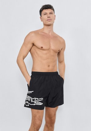 Bañador corto negro hecho de un material ligero, con un gráfico blanco de un cocodrilo en la pierna izquierda y una cintura elástica con bolsillos.