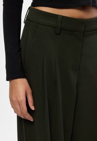 Το αριστερό χέρι resting δίπλα σε σκούρο πρ�άσινο πλατύ παντελόνι και μια μαύρη μακριά μπλούζα με κοντό μανίκι, ορατό από την περιοχή της μέσης μέχρι το μηρό.