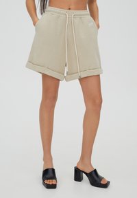 Shorts en coton beige avec revers, taille élastique, cordon de serrage et poches latérales. Présente une broderie discrète du logo blanc.