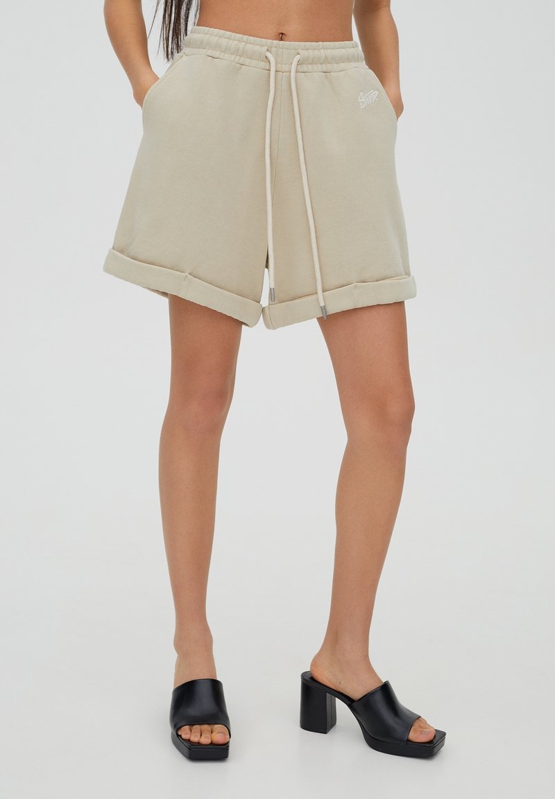 Shorts en coton beige avec revers, taille élastique, cordon de serrage et poches latérales. Présente une broderie discrète du logo blanc.