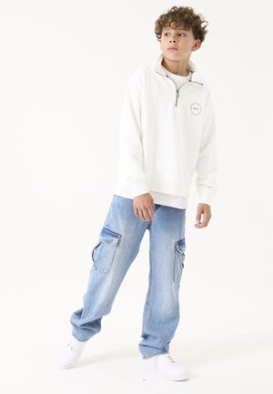 Sudadera blanca con cierre y logo en el pecho, combinada con pantalones cargo azul claro que tienen dos bolsillos laterales y zapatillas blancas.