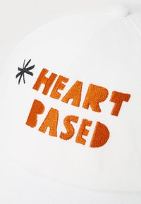 Bijela kapa s narančastim izvezenim tekstom "HEART BASED" i malim crnim zvjezdastim akcentom. Pamuk s oblikovanim oblikom.