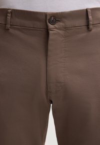 Pantaloni di cotone marroni con chiusura a bottone, tasche laterali e trama liscia. Presentano una vestibilità slim e un design a gamba dritta.