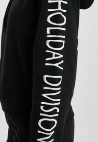 Čierna mikina s kapucňou vyrobená z bavlny, s voľným strihom. Na rukáve má biele písmo s nápisom "HOLIDAY DIVISION." Sťahovacia kapucňa.