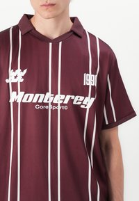 Burgunderfarget kortermet jersey med hvite vertikale striper. Har kragekrage, frontlogo "Monterey" og året "1990."