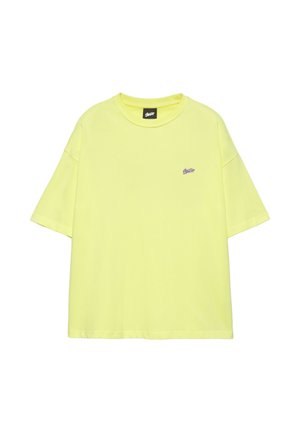 Jeune homme portant un t-shirt jaune, un short en jean jusqu’aux genoux, des chaussettes blanches et des baskets noires et blanches, debout contre un mur blanc uni.