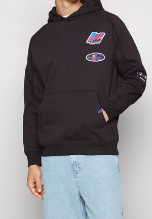 Person iført sort hættetrøje med farverige grafikpletter og lyseblå jeans, hånd delvist i forlomme.