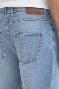 Vaqueros de mezclilla azul claro con una textura suave, dos bolsillos traseros y un parche de cuero marrón en la cintura con la etiqueta "!SOLID".