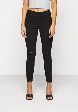 Femme portant un jean skinny noir taille haute déchiré avec des ourlets effilochés, associé à un haut court gris et des talons noirs ouverts à l'avant.