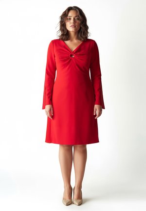 Rotes Kleid mit langen Ärmeln, V-Ausschnitt und einer Knotendetail am Brustbereich. Glatter Stoff, knielang, kombiniert mit beigen Absätzen.