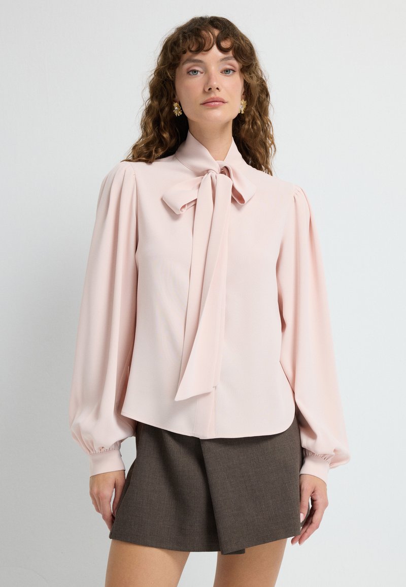 Blusa rosa chiaro con colletto a fiocco, maniche puff lunghe e una texture liscia, abbinata a una gonna marrone con un design sartoriale.