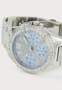 Montre argentée avec un cadran bleu, des accents en cristal et trois sous-cadrans. Dispose d'un bracelet en acier inoxydable et d'un fond texturé sur le cadran.