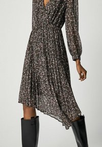 Svart blommig klänning med långa ballongsärmar, markerad midja, flödande plisserad kjol och asymmetrisk fåll. Matchad med knähöga svarta stövlar.