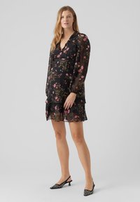 Vestido negro con estampado de flores, mangas largas transparentes, falda en capas y escote en V. Presenta patrones de flores en tonos rosa y verde, complementados con zapatos de tacón negros.