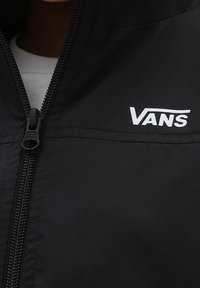 Czarna kurtka na zamek wykonana z gładkiej tkaniny, z białym logo Vans na lewej piersi oraz zamkiem błyskawicznym z przodu.