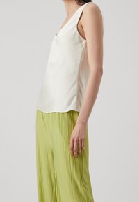 Weißes Satin-Oberteil mit V-Ausschnitt, ärmellos und glatter Textur. Kombiniert mit plissierten, hellgrünen Hosen, die vertikale Linien zeigen.