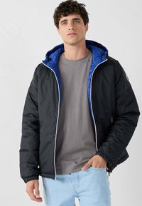 Giacca a vento nera con cappuccio trapuntato blu, dotata di zip frontale e polsini elasticizzati. Indossata sopra una camicia grigia e jeans chiari.