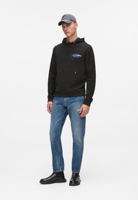 Sudadera negra con bolsillo frontal y logo, jeans azul claro con detalles desgastados y botas negras hasta el tobillo. Gorra gris completa el atuendo.