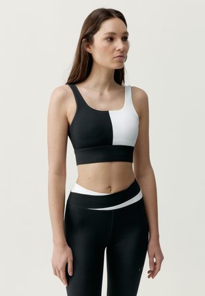 Kvinde iført en sort og hvid sports-bh med farveblokering og matchende højtaljede leggings, stående med højre arm afslappet og kigger mod højre.