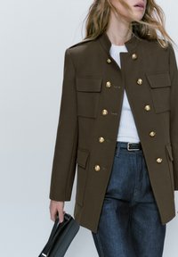 Veste militaire marron, boutons dorés, col haut, quatre poches à l'avant, portée sur un haut blanc avec un jean bleu, tenant un sac noir.