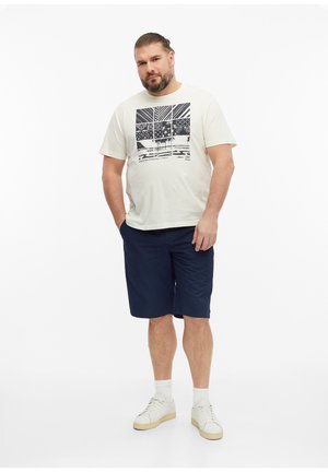 Man met baard die een wit grafisch T-shirt draagt, marineblauwe shorts, witte sokken en witte sneakers, staat met één hand in zijn zak.
