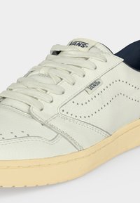 Λευκό αθλητικό παπούτσι Vans με κρεμ σόλα, διάτρητο μοτίβο στα πλευρά, λευκά κορδόνια και λογότυπα Vans στη γλώσσα και στην πλαϊνή ετικέτα.