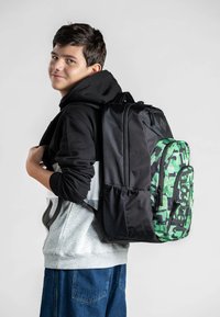 Zaino mimetico nero e verde con compartimenti con zip, superfici texture e schienale imbottito, indossato da una persona in abbigliamento casual.