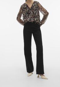 Blusa fluida floreale con maniche lungheTrasparenti, abbinata a pantaloni neri a vita alta e ampia, e scarpe a punta di colore chiaro.