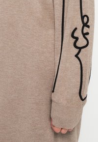 Pull en tricot beige avec un motif de dessin linéaire noir le long des manches, doté d'une texture côtelée et de poignets longs pour plus de couverture.