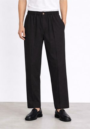 Mężczyzna noszący czarne, dopasowane spodnie z elastycznym pasem i czarne skórzane wsuwane buty, stojący na szarej podłodze.