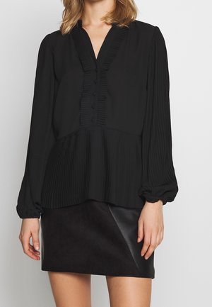 Kobieta w czarnej plisowanej bluzce z długim rękawem i guzikami oraz czarnej skórzanej mini spódnicy na jednolitym tle.