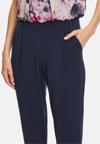 Femme portant un pantalon taille haute bleu marine avec des plis et des poches, associé à un chemisier floral rentré.