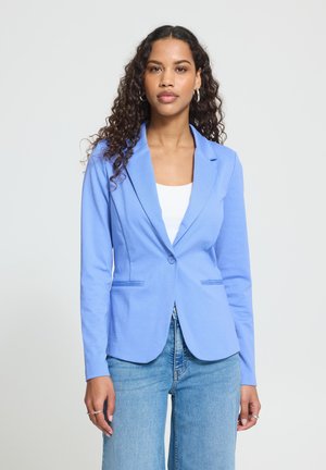 Femme aux longs cheveux bouclés portant un blazer bleu clair, un top blanc et un jean bleu, debout devant un fond uni clair.
