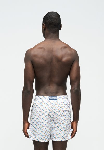 Άνδρας που φαίνεται από πίσω, φοράει λευκά μαγιό σορτς με πολύχρωμα σχέδια αστεριών και ετικέτα Vilebrequin στη μέση.