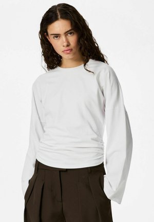 Jeune femme aux longs cheveux bouclés portant une chemise blanche à manches longues et un pantalon plissé taille haute marron foncé, debout devant un fond blanc.