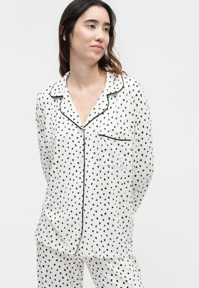 Νεαρή γυναίκα φοράει λευκό σετ πιτζάμες με μικρά μαύρα σχέδια καρδιών και μαύρη επένδυση, στέκεται με τα χέρια πίσω από την πλάτη.