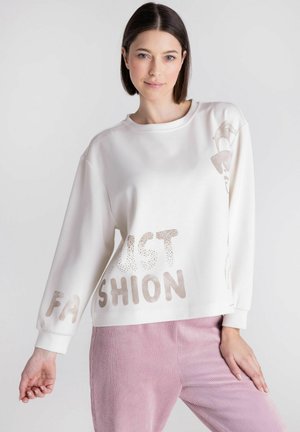 Vrouw met steil donker haar, gehuld in een witte trui met de tekst "FAST FASHION" en roze ribfluwelen broek tegen een effen lichte achtergrond.