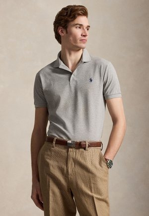 Ung mand iført lysegrå polo shirt og beige bukser med brun læderbælte, stående med den ene hånd i lommen, kiggende mod højre.