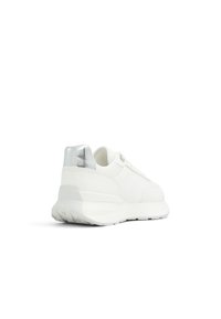 Zapatilla baja blanca con suela gruesa y tab de talón metálico plateado, mostrada desde un ángulo trasero izquierdo sobre fondo blanco.