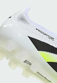 White soccer cleat with textured upper, black accents, prominent branding, and transparent studded sole. Features a padded collar.