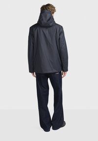 Pánská černá voděodolná bunda s velkou kapucí, s hladkým povrchem a uvolněným střihem, kombinovaná s tmavými džíny.