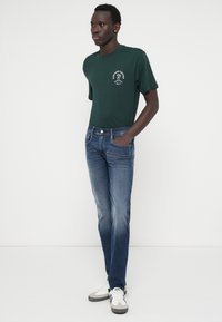 Σκούρο πράσινο βαμβακερό μπλουζάκι με λογότυπο στο στήθος, σε συνδυασμό με μπλε τζιν με ξεθωριασμένα χρώματα και κανονική γραμμή, ολοκληρωμένο με λευκά αθλητικά παπούτσια.